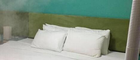 A bed with white linens and pillows, a folded towel on the bed, and a white ceiling vent emitting steam.