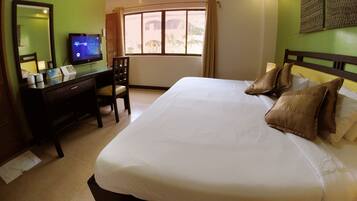A hotel room with a large bed, a desk with a computer, a chair, a window with curtains, and a wall-mounted air conditioner.