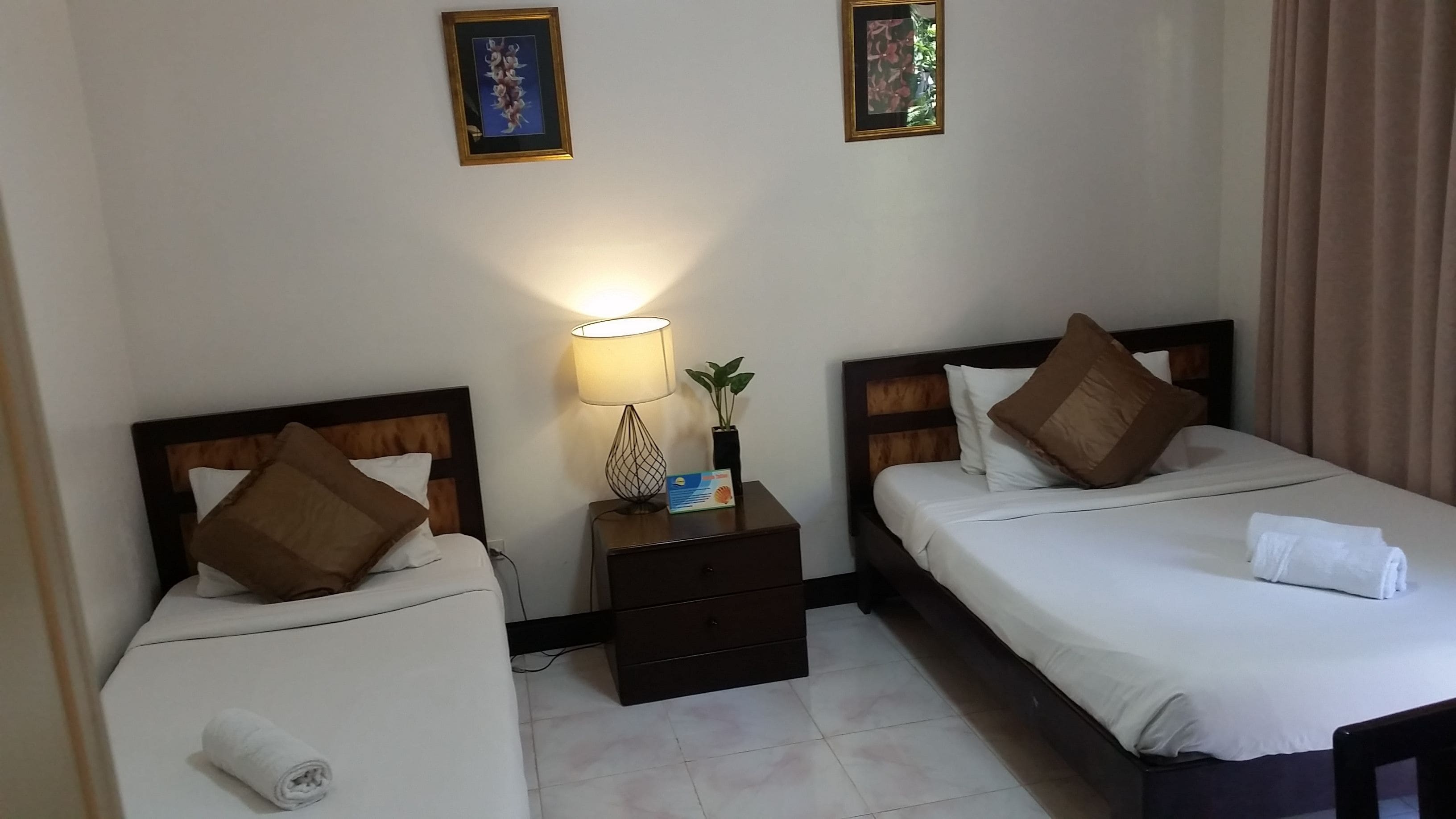 Een hotelkamer met twee enkele bedden, een nachtkastje met een lamp, een kleine plant en een ingelijste foto aan de muur.