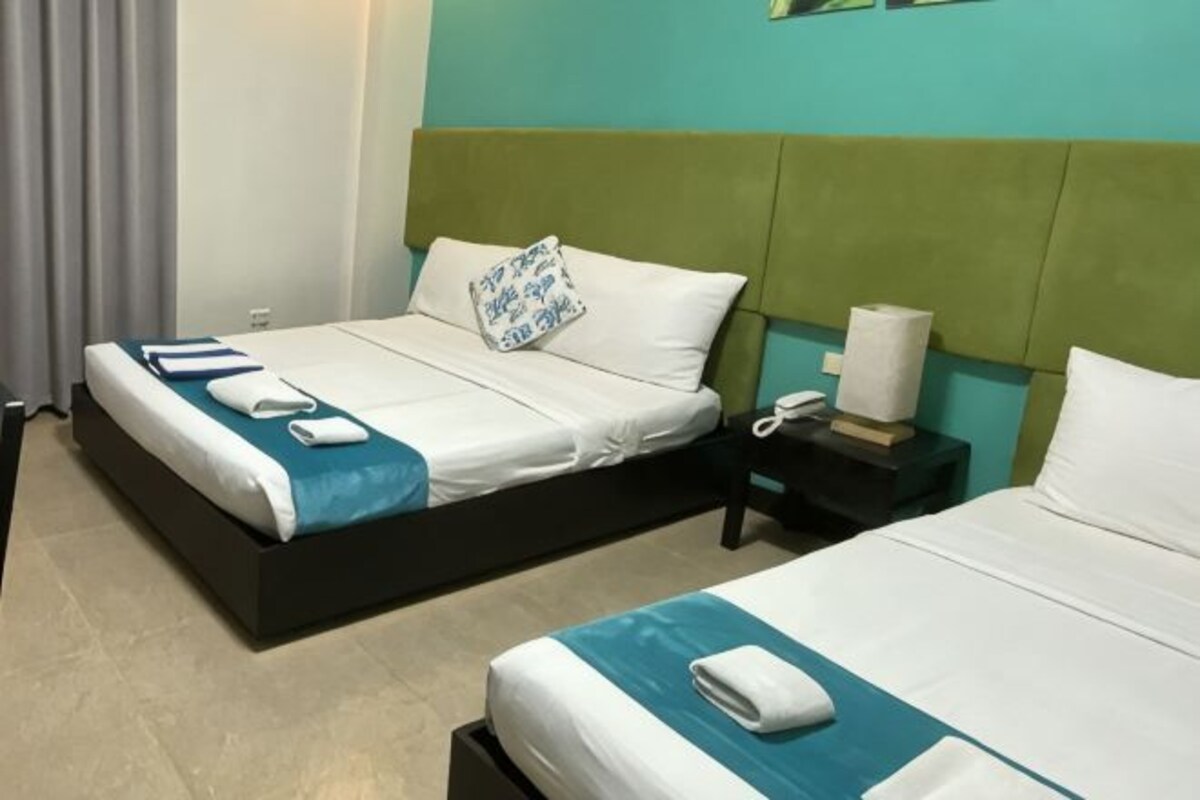 Habitación de hotel con dos camas, cabeceros verdes, almohadas y toallas blancas en cada cama, además de un teléfono blanco.