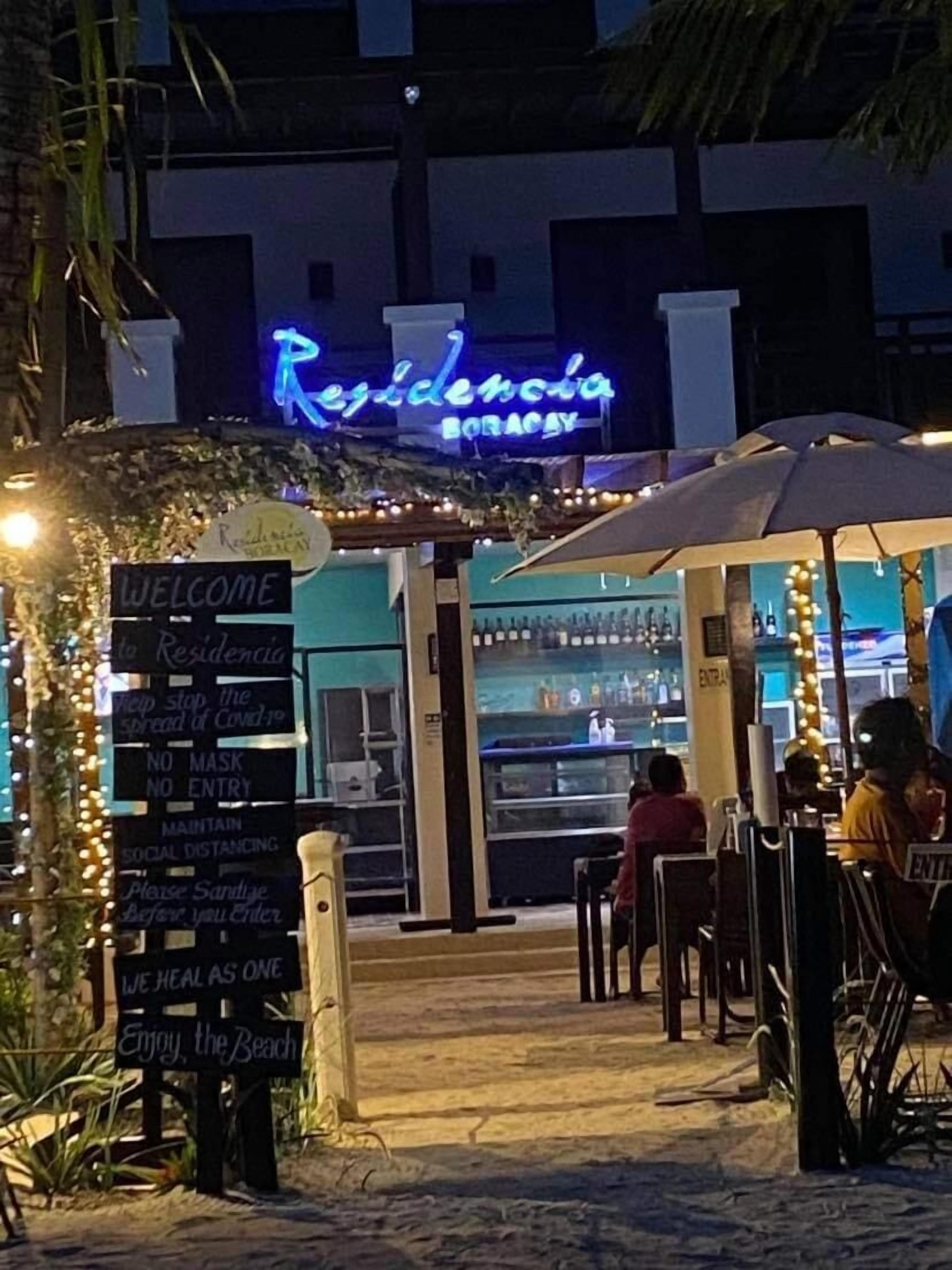A beachfront restaurant with outdoor seating, a neon sign reading "Residencia Boracay", and a sign listing COVID-19 safety measures.