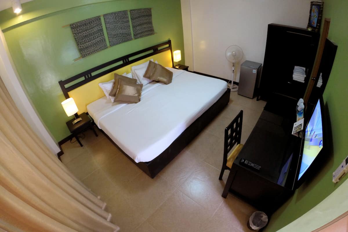 Quarto de hotel com cama, abajures, escrivaninha com computador, televisão e pequena geladeira.