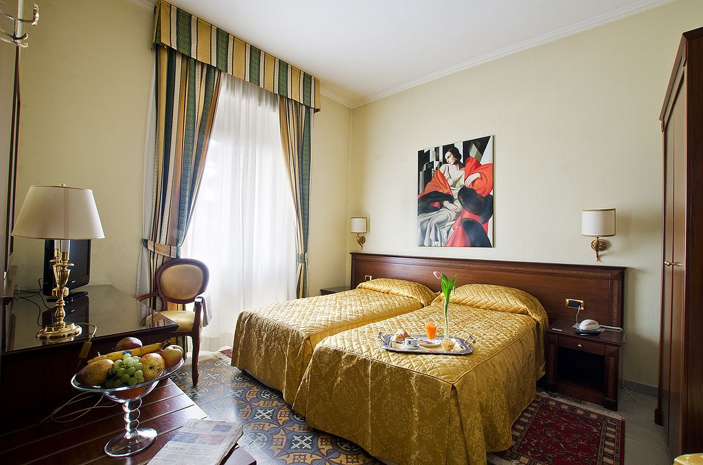 Een hotelkamer met twee bedden, een eettafel met fruit, een stoel, een schilderij aan de muur en een raam met gordijnen.