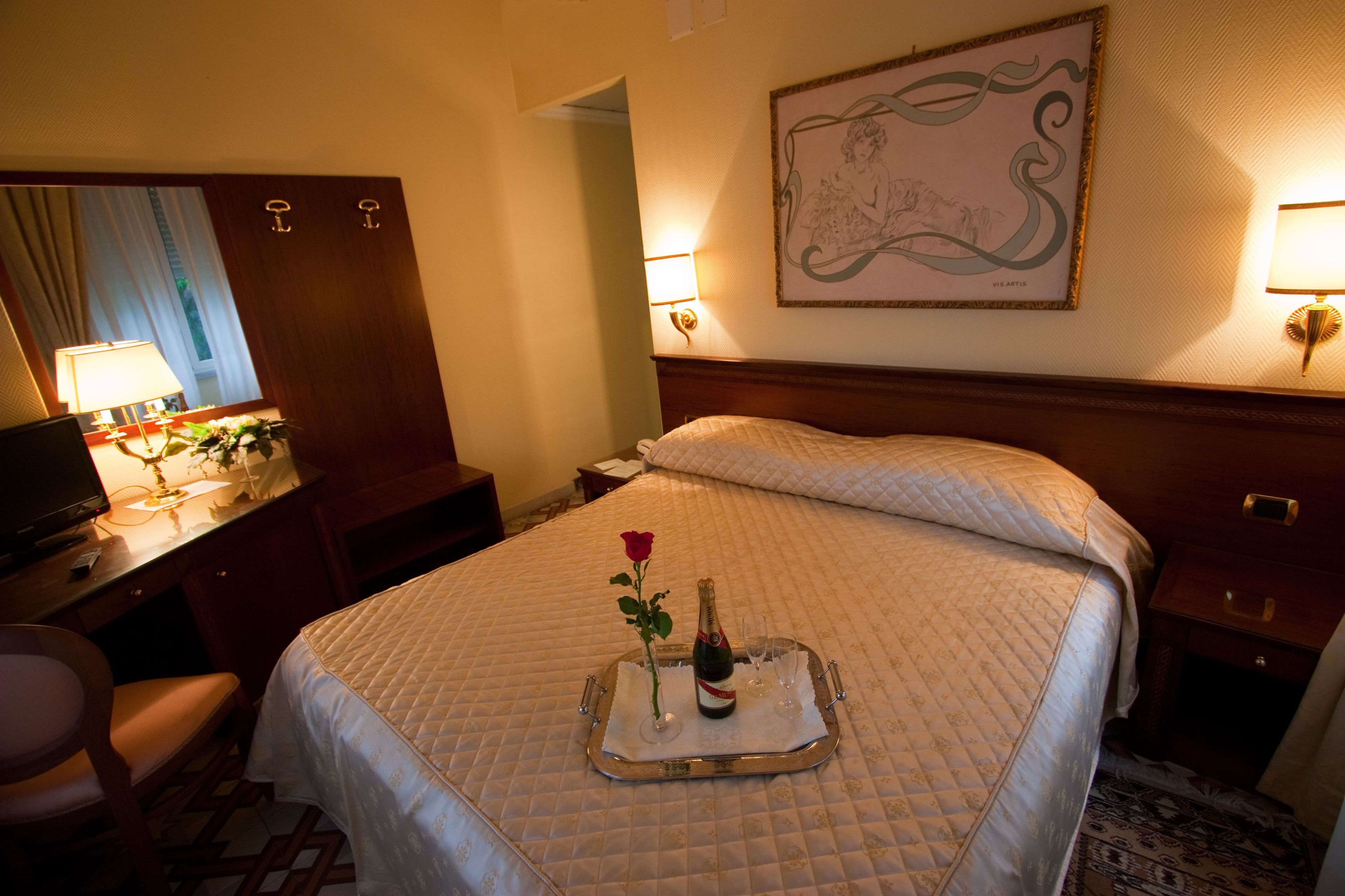 A hotel room with a bed, a desk with a lamp, a chair, a framed artwork on the wall, and a tray with a rose and champagne.