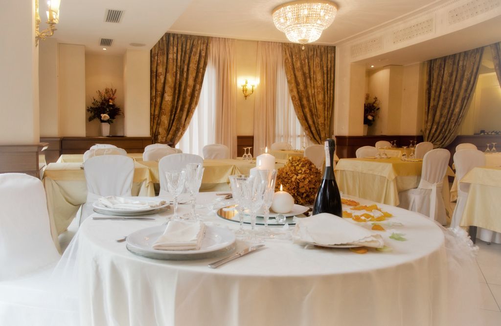 A formal dining room with round tables set for a meal, featuring white tablecloths, chairs, and a chandelier.