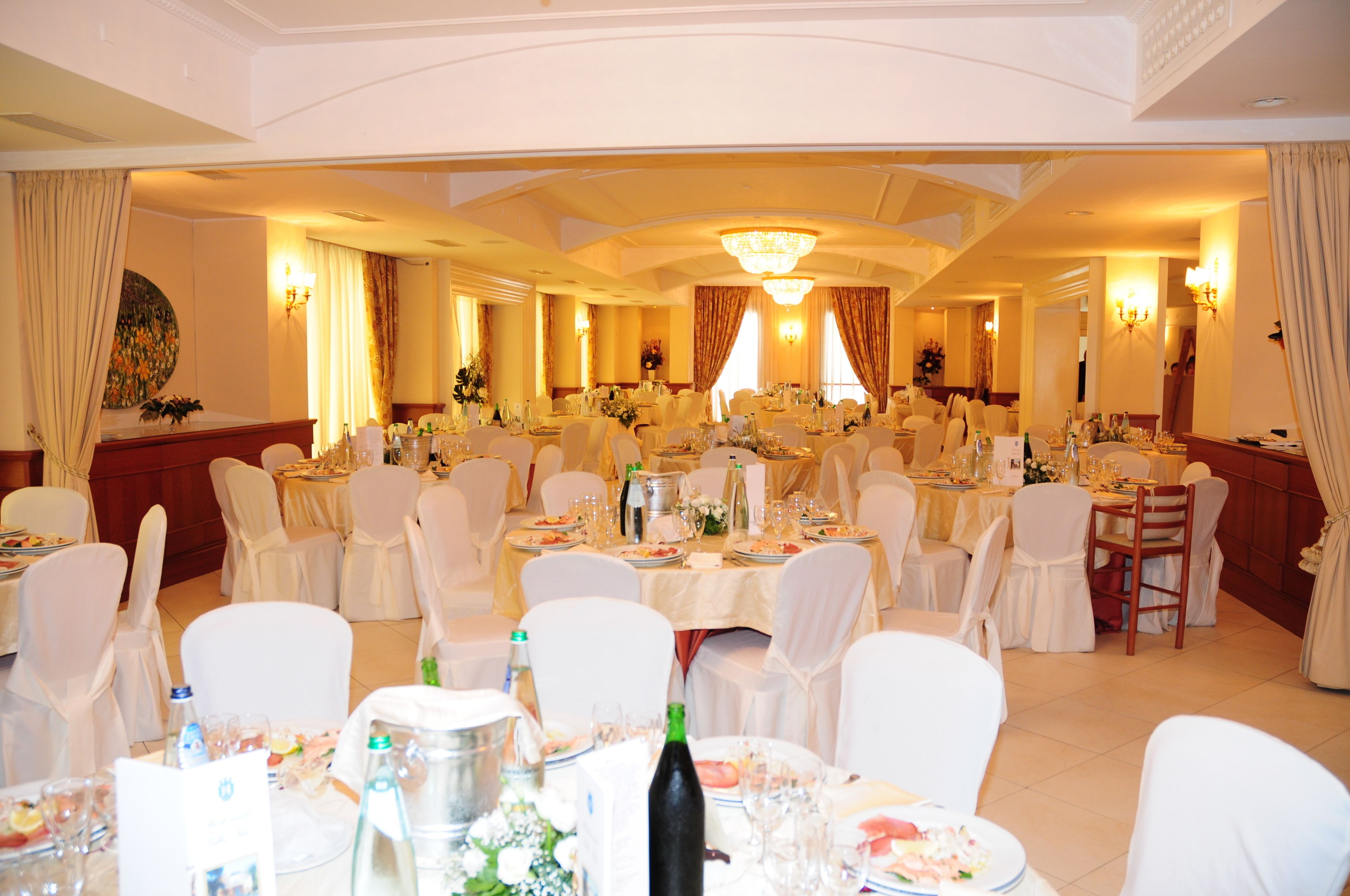 A banquet hall with round tables set for dining, white chairs, and chandeliers.