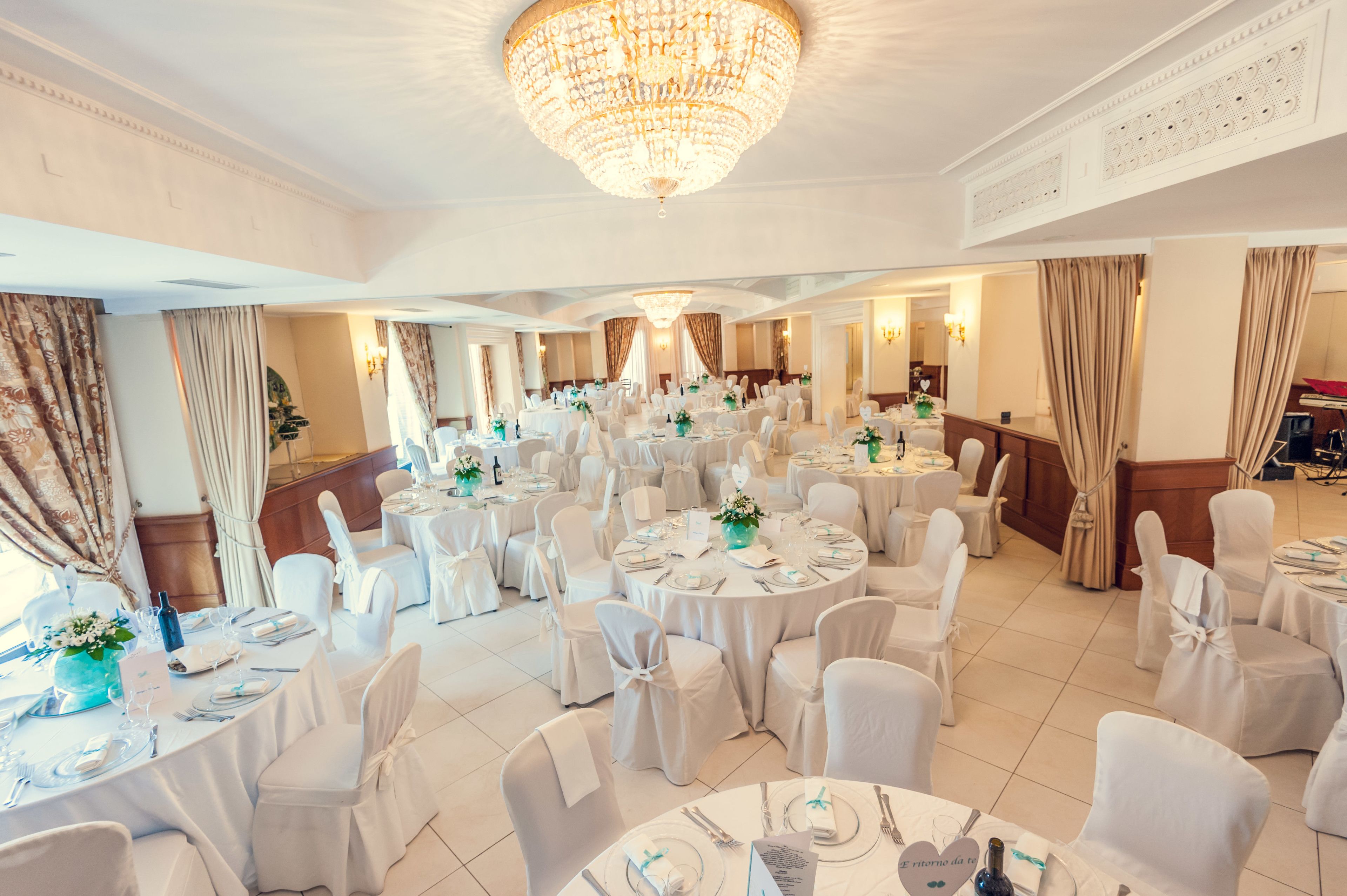 A banquet hall with round tables, white chairs, and chandeliers.