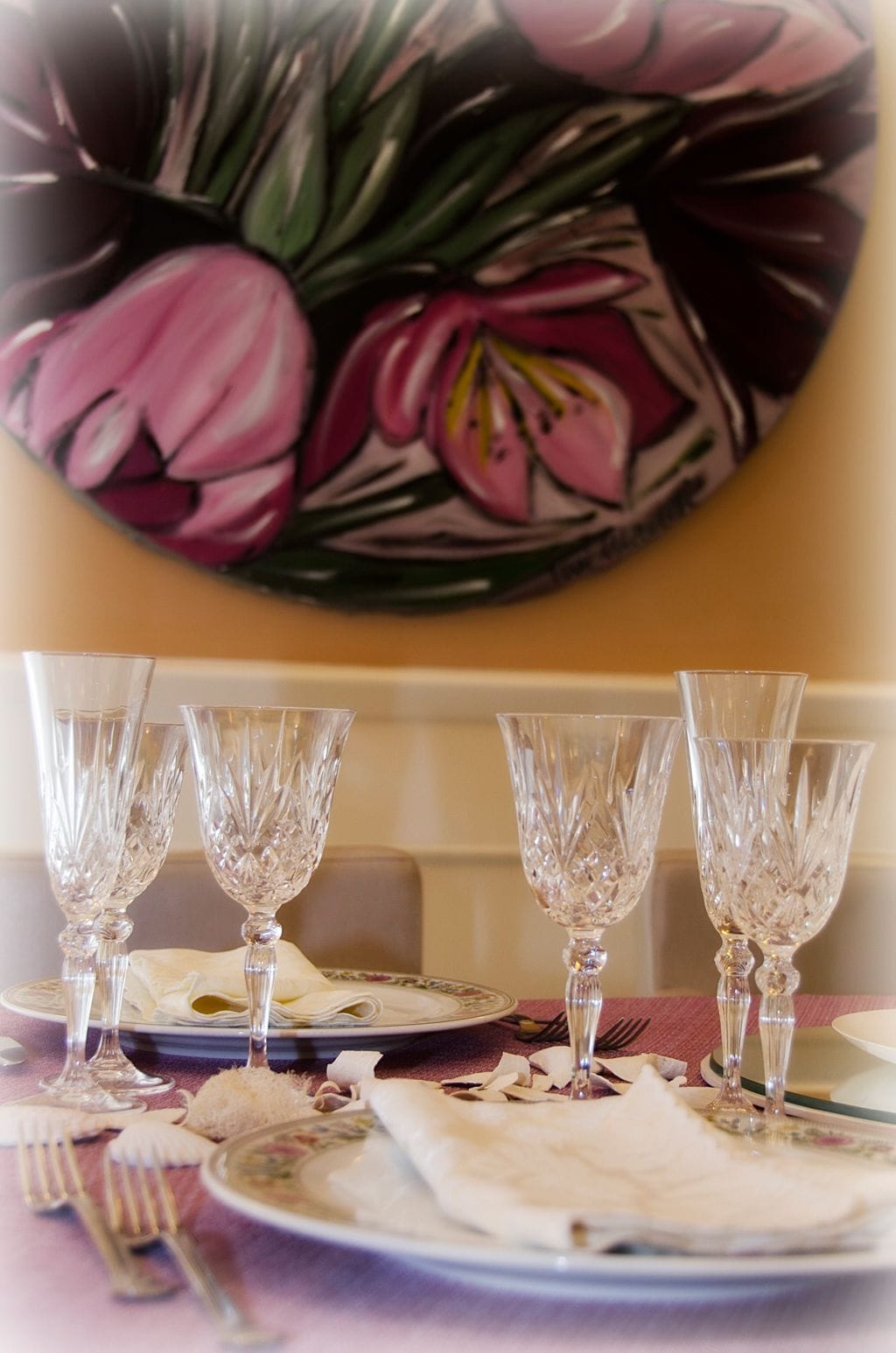 Una mesa de comedor dispuesta con cristalería y platos, con un cuadro floral en la pared.