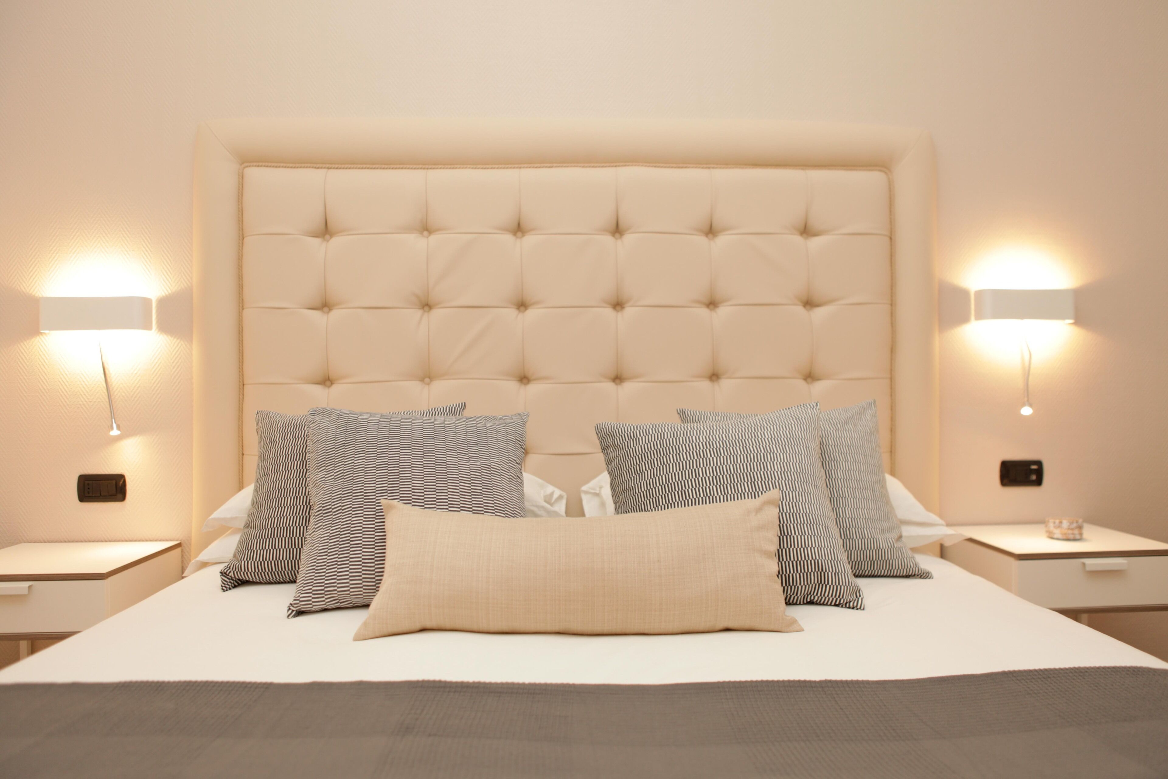 Una cama bien hecha con un cabecero capitonado, varios almohadones y mesitas de noche con lámparas fijadas a la pared.
