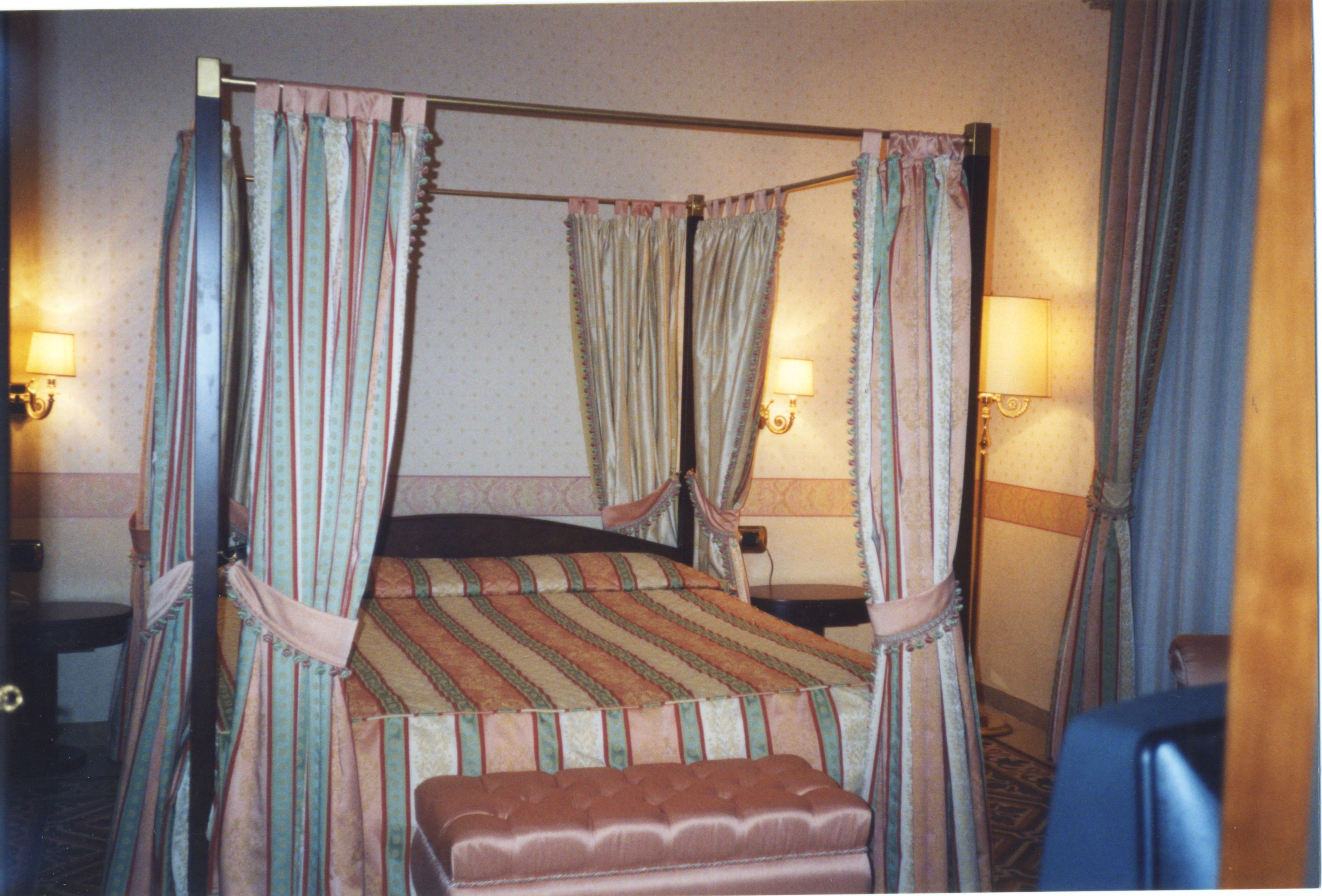 Una cama con dosel, ropa de cama a rayas, dos lámparas de noche y un banco al pie de la cama.