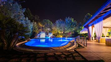 Piscina illuminata di notte, circondata da alberi e con un edificio con tende bianche.
