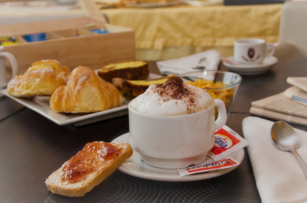 A breakfast setting with croissants, a cup of coffee with whipped cream, and a slice of toast with jam.