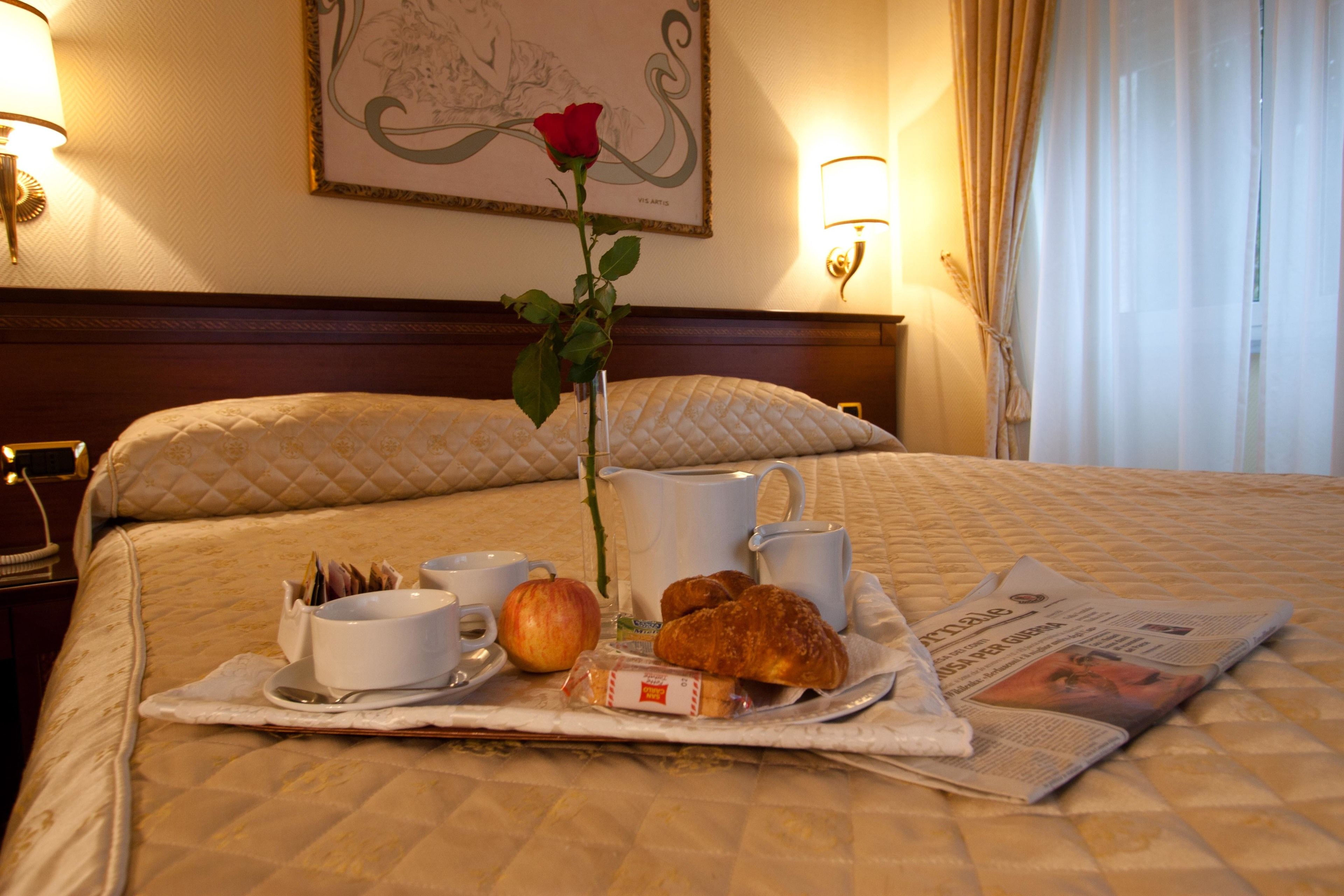 Un plateau de petit-déjeuner soigneusement préparé est posé sur le lit, qui est recouvert d’une courtepointe matelassée. On y trouve un vase avec une seule rose, un journal, un croissant, une tasse et une assiette avec une cuillère.