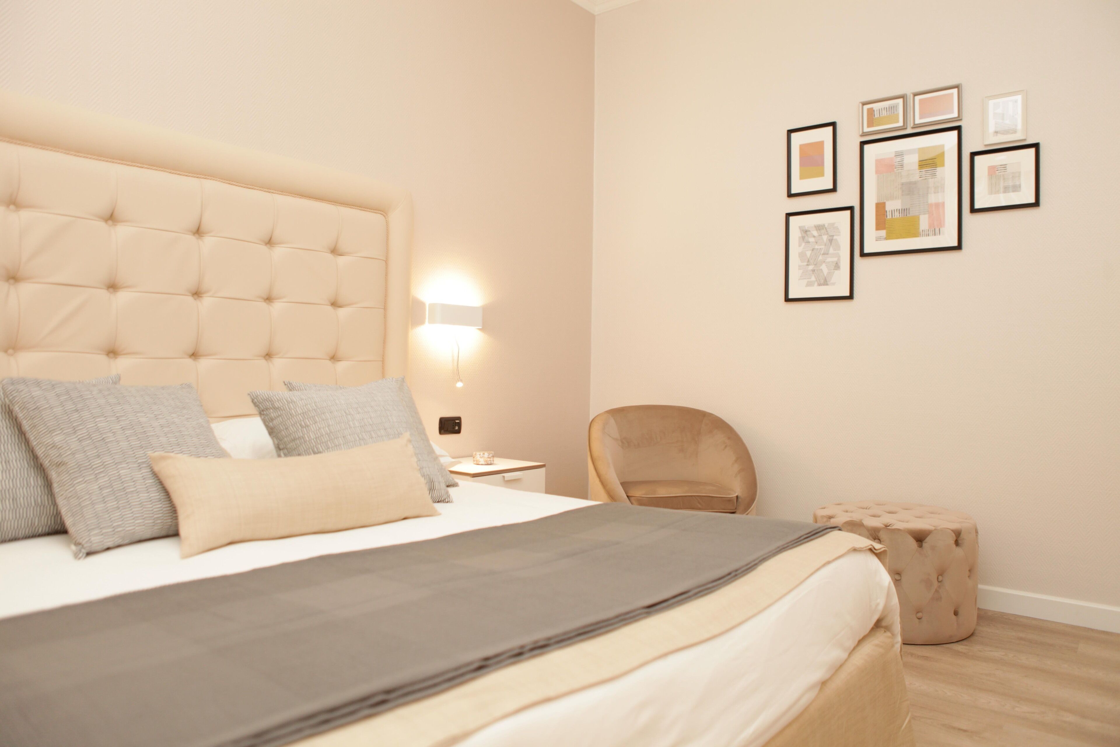 Ein Schlafzimmer mit einer gepolsterten Kopfteile, einem Bett mit grau-beiger Bettwäsche, einem Nachttisch mit Lampe, einem Sessel und einem kleinen Hocker.