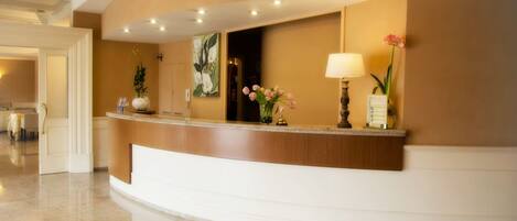 A hotel reception area with a curved reception desk, a large mirror, a wall-mounted lamp, and a vase with flowers.