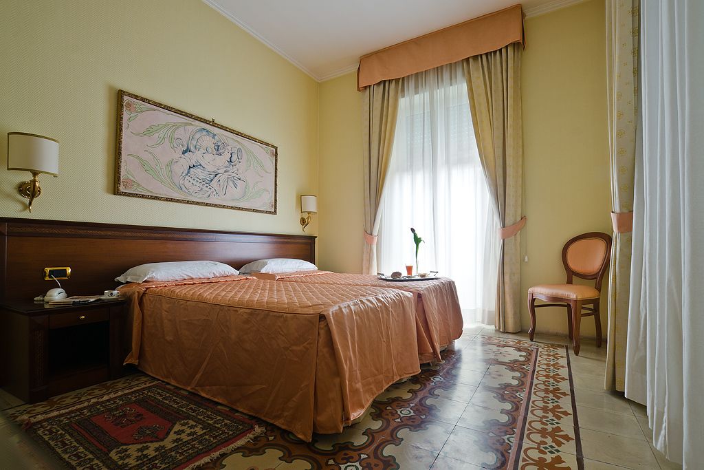 A hotel room with a large bed, two bedside tables, a chair, a painting on the wall, and a window with curtains.