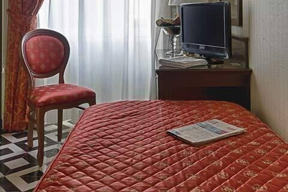 Une chambre d’hôtel avec un lit, une chaise, un bureau avec une télévision et une fenêtre avec des rideaux.