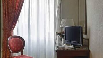 Habitación de hotel con cama, una silla, escritorio con televisor y ventana con cortinas.