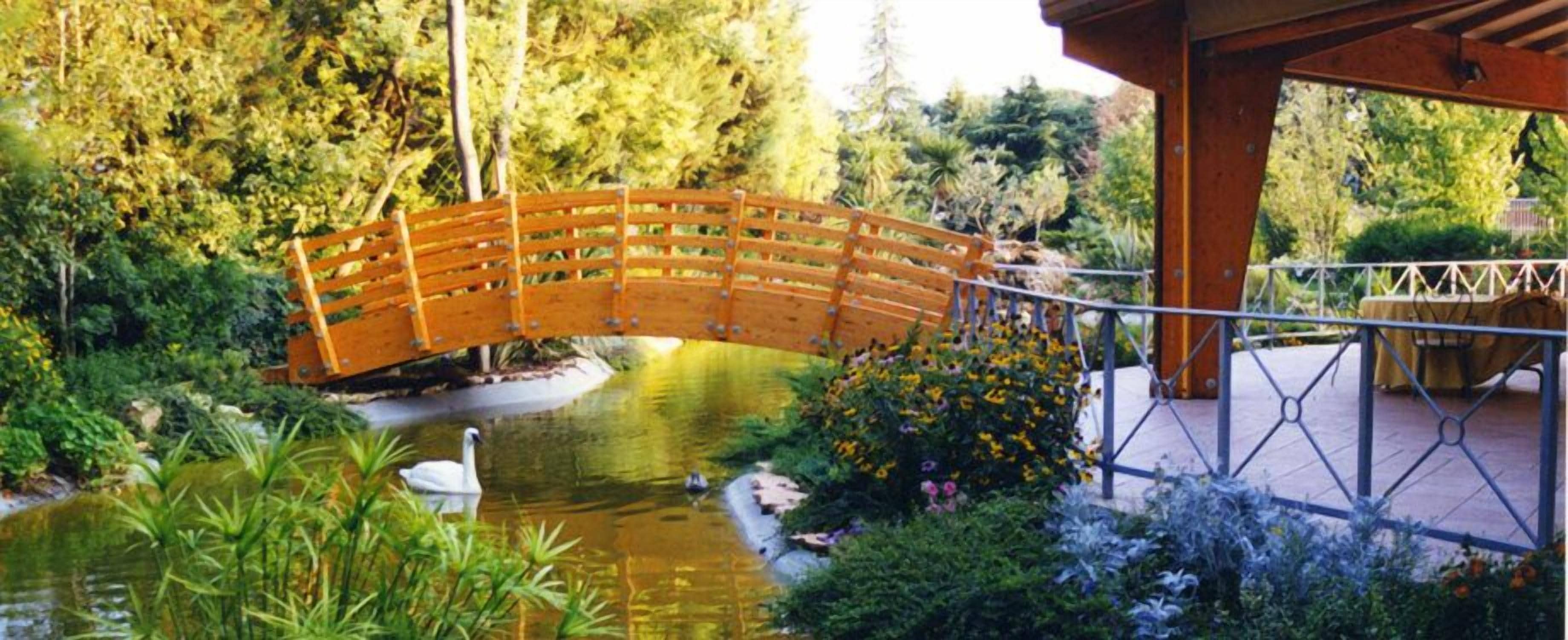 Un puente de madera sobre un estanque con cisnes, rodeado de abundante vegetación y una zona de estar cubierta.