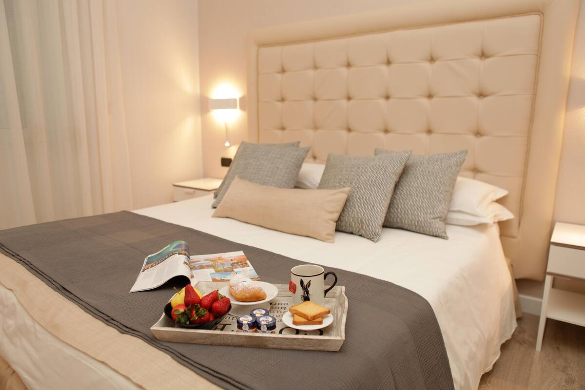 Un lit bien fait, avec un plateau contenant divers éléments pour le petit-déjeuner, notamment des fruits, des viennoiseries et une tasse, dans une chambre d’hôtel moderne.