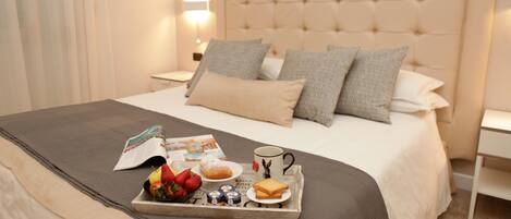 Una cama bien tendida con una bandeja de desayuno que incluye frutas, pasteles y una taza, en una habitación de hotel moderna.