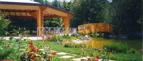 Un pabellón con techo de madera y baranda blanca, rodeado por un jardín con un estanque y un puente de madera.