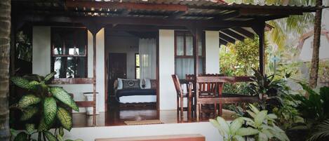 A covered porch with a wooden bench and table, surrounded by tropical plants.