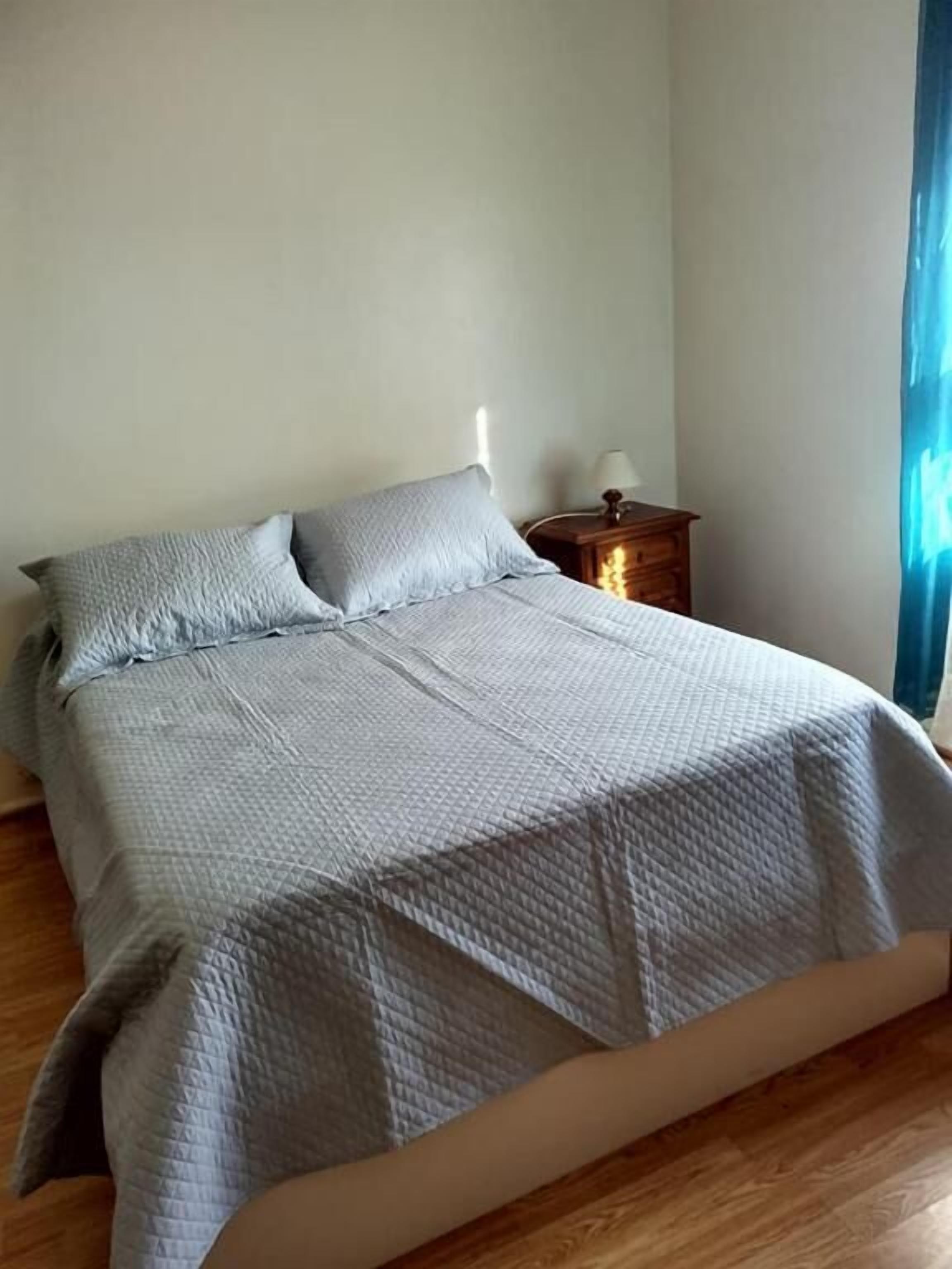 Una cama bien hecha con cubrecama acolchado, una mesita de noche de madera con una lámpara y una ventana con cortinas.