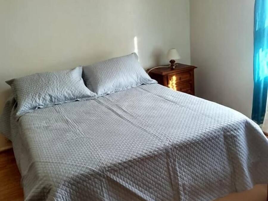 Un letto rifatto con copriletto trapuntato, un comodino in legno con una lampada e una finestra con tende.