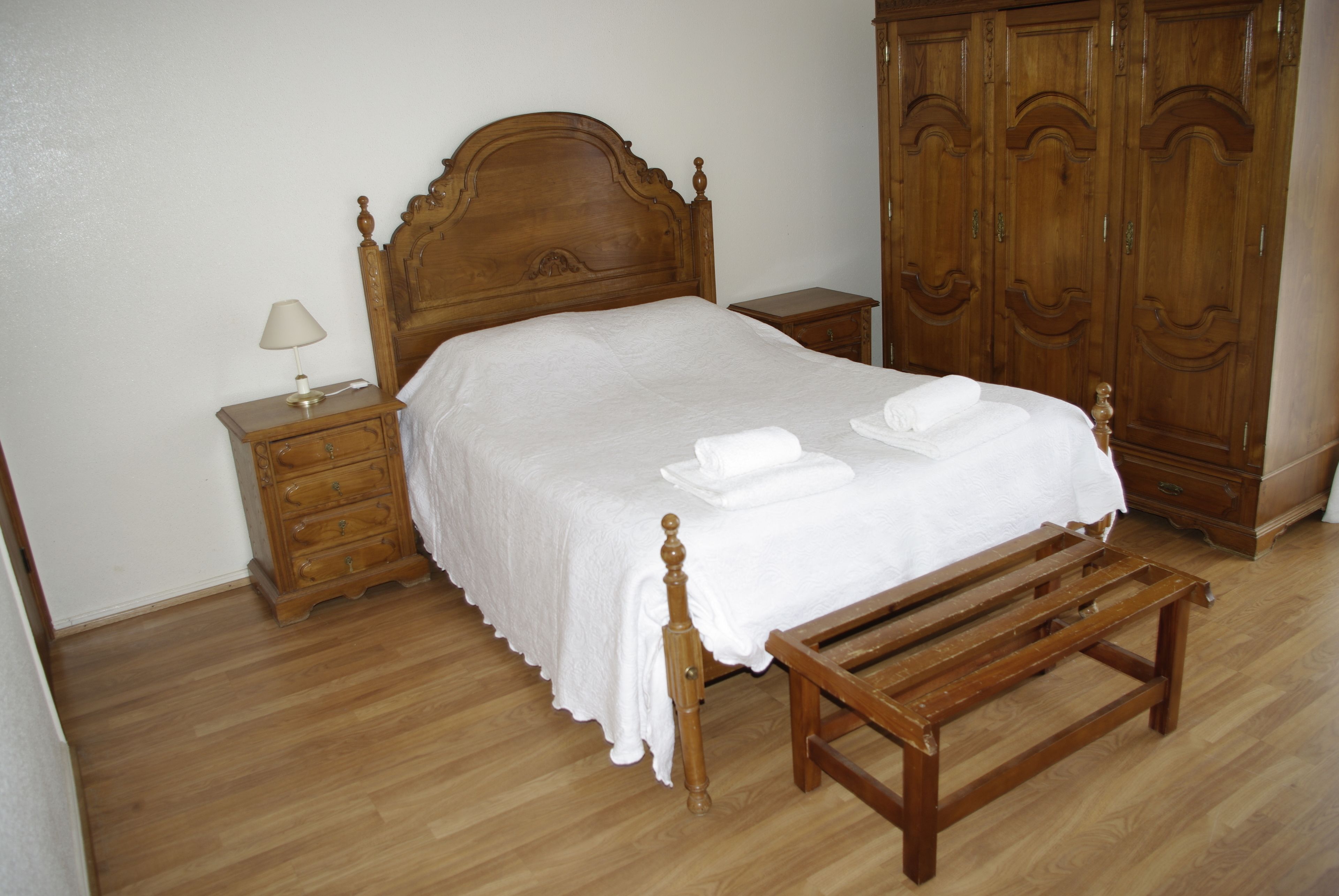 Una cama bien hecha con cabecera de madera, una mesita de noche de madera con lámpara y un banco de madera.