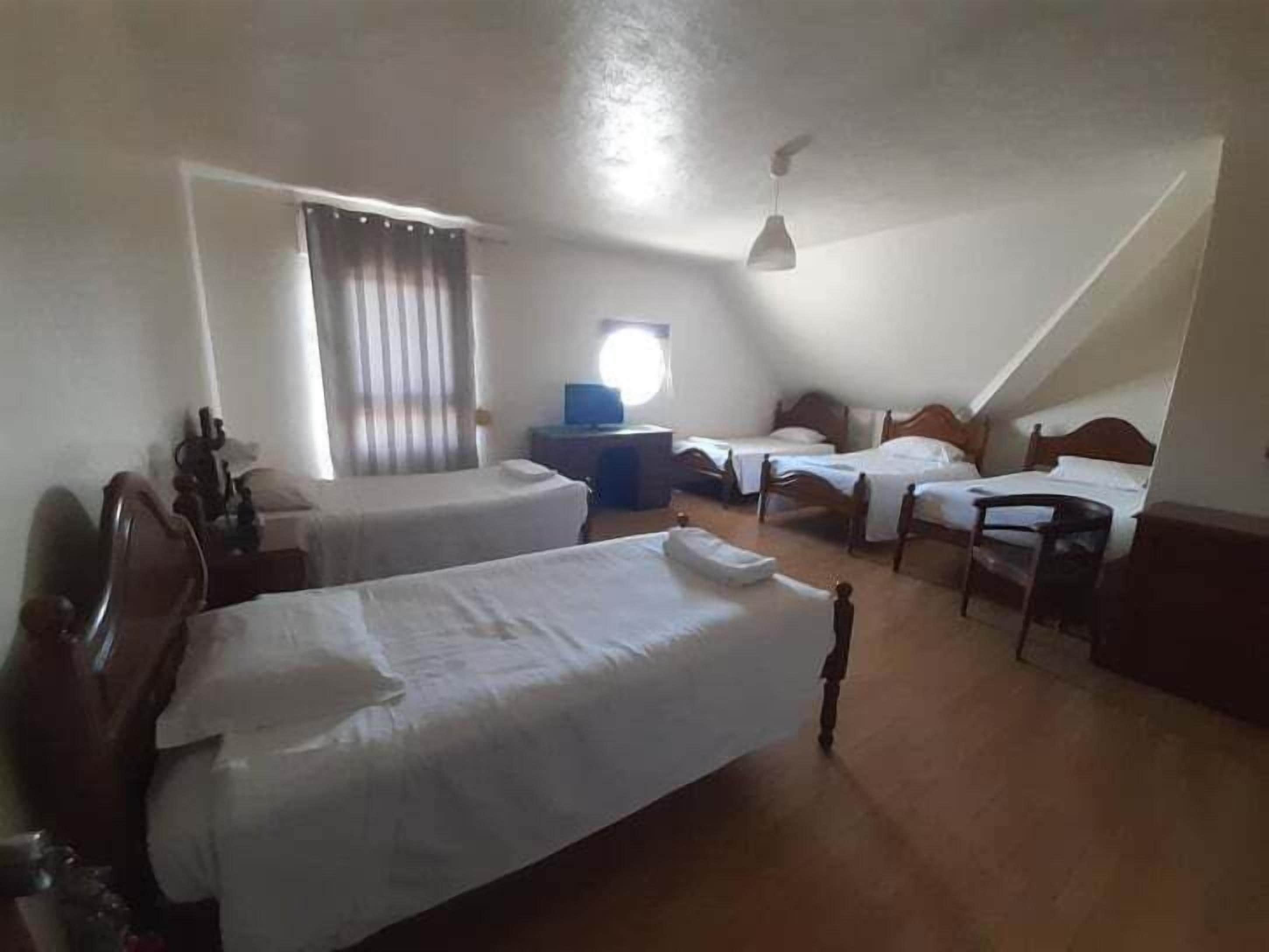 Quarto com duas camas de solteiro, uma escrivaninha, uma cadeira, janela com cortinas e ventoinha no teto.