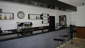 Zona de bar con una cafetera, estantes con botellas, un reloj y taburetes de bar.