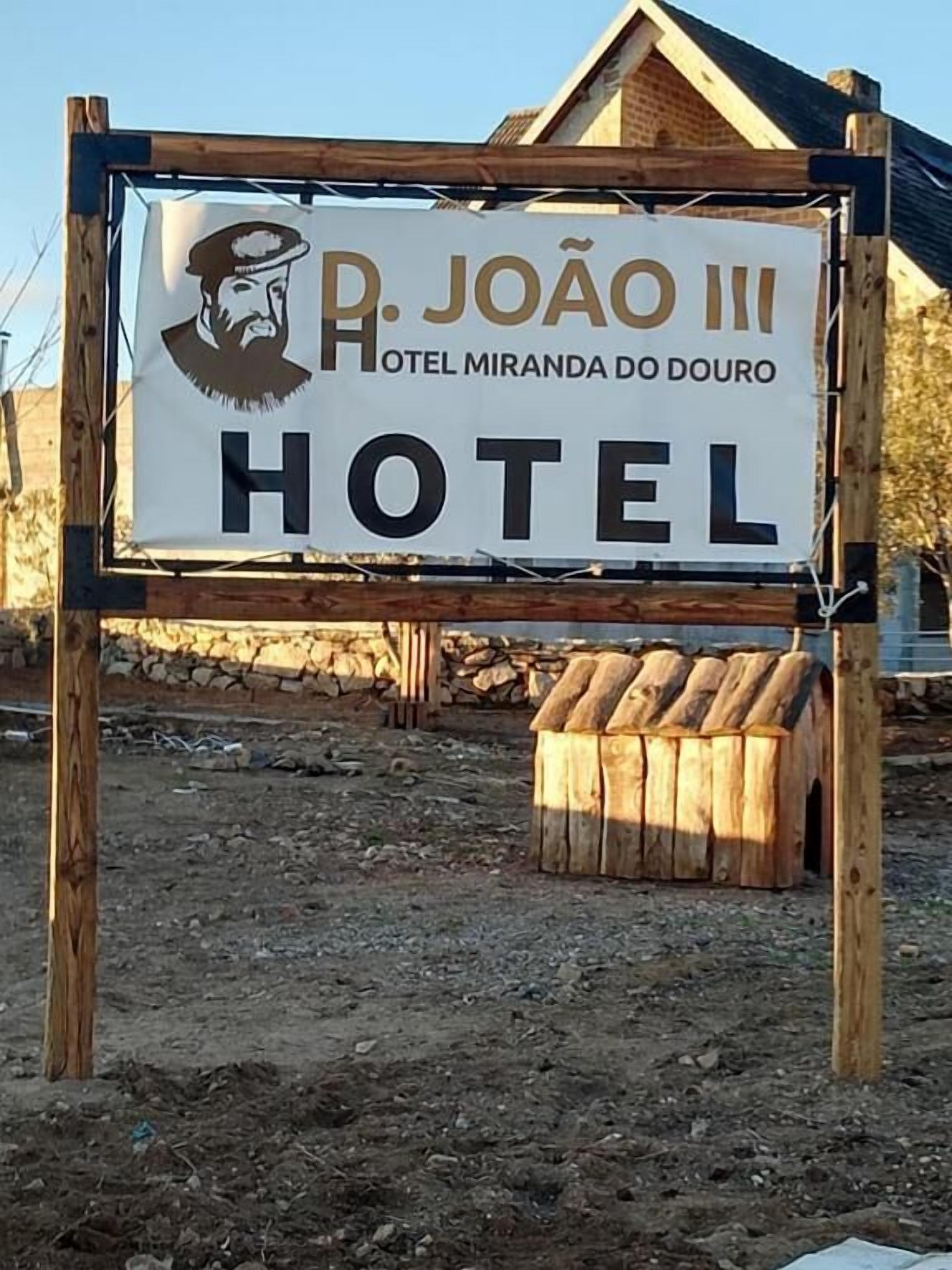 Ein Schild für das 'R. João III Hotel Miranda do Douro' mit einer rustikalen Holzkonstruktion und einer dahinterliegenden Steinmauer.