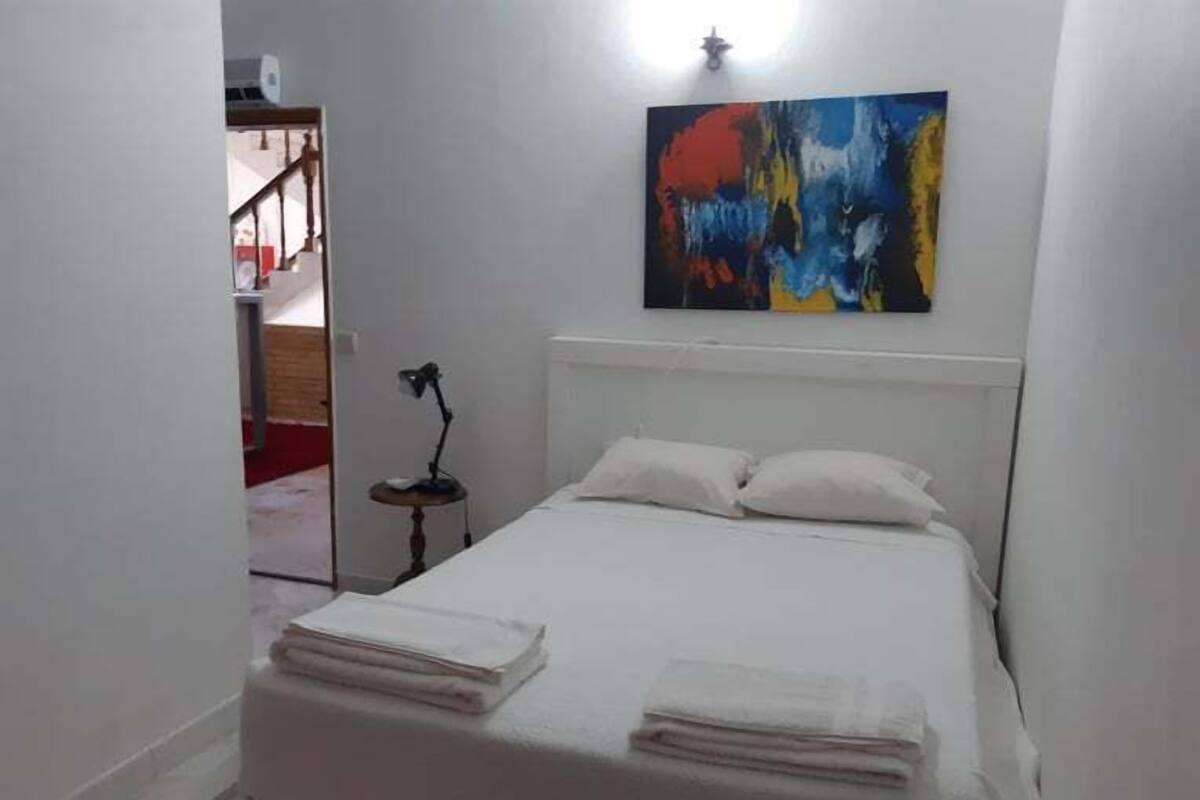 Een slaapkamer met een bed, kussens, een bureau met een lamp en een schilderij aan de muur.