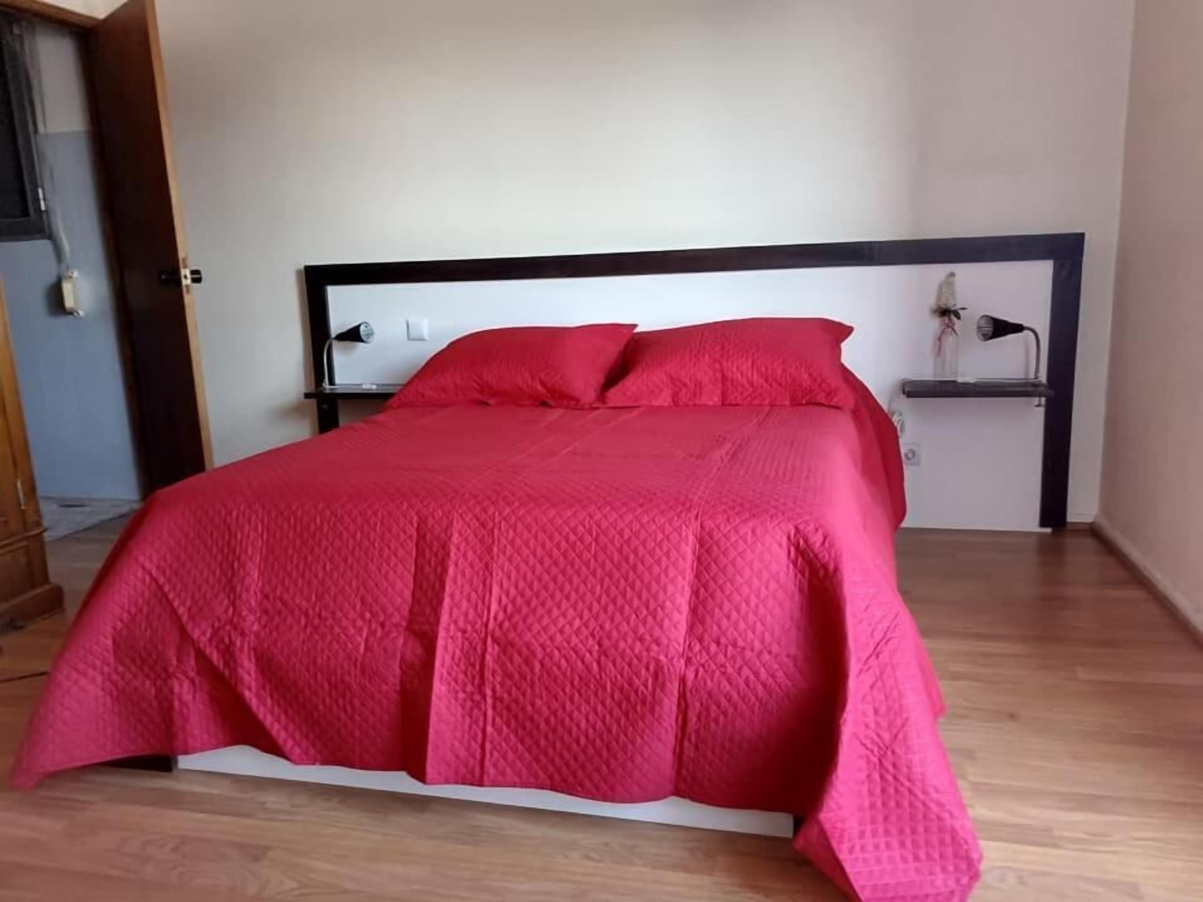 A bed with a red quilted cover, a wooden headboard, and a bedside table with a lamp.