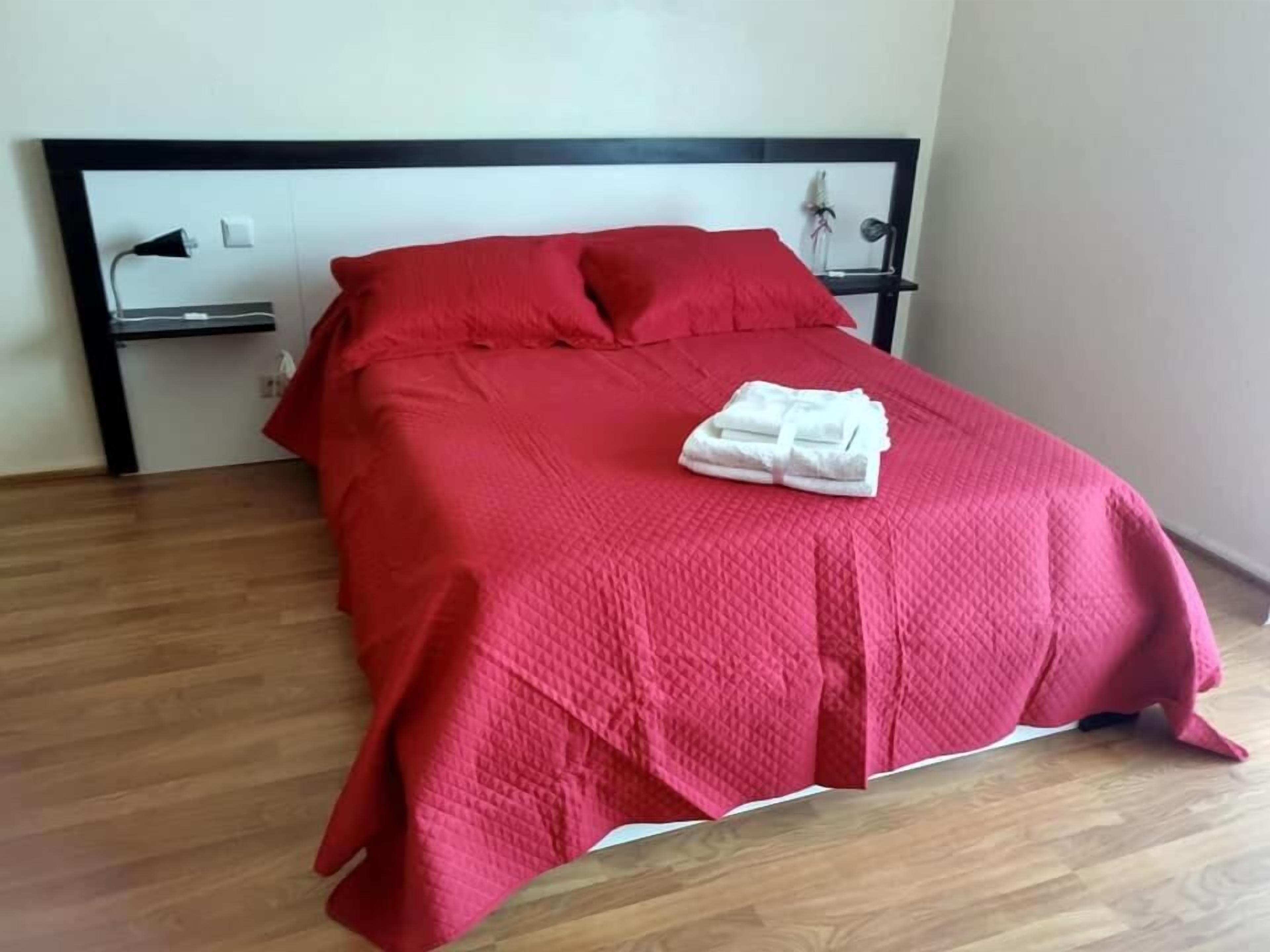 Een netjes opgemaakt bed met een rode sprei, witte handdoeken, een zwart hoofdeinde en een houten vloer.