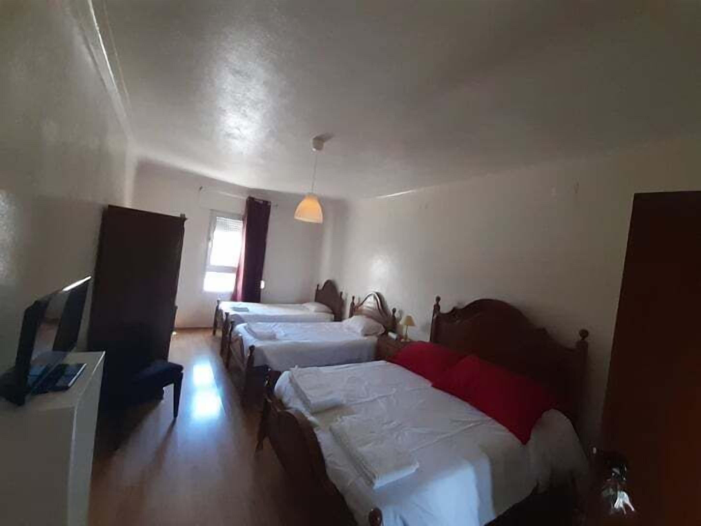 A room with three single beds, a desk, and a chair. There is a window with curtains and a hanging light fixture.