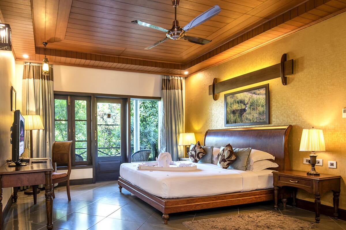 A spacious bedroom with a large bed, wooden ceiling, and a ceiling fan. There is a desk with a chair and a television.