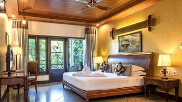 A spacious bedroom with a large bed, wooden ceiling, and a ceiling fan. There is a desk with a chair and a television.