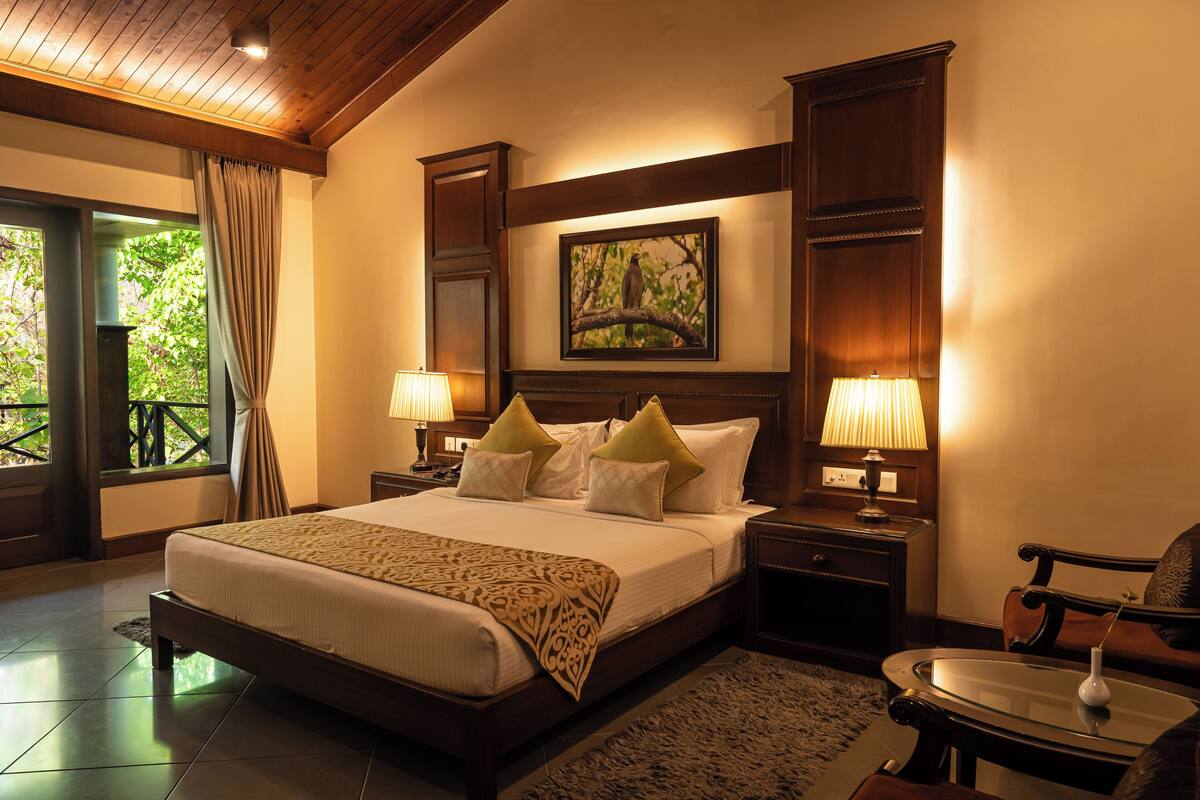 A spacious bedroom with a large bed, wooden headboard, two bedside tables with lamps, a nightstand, and a view of greenery through a window.