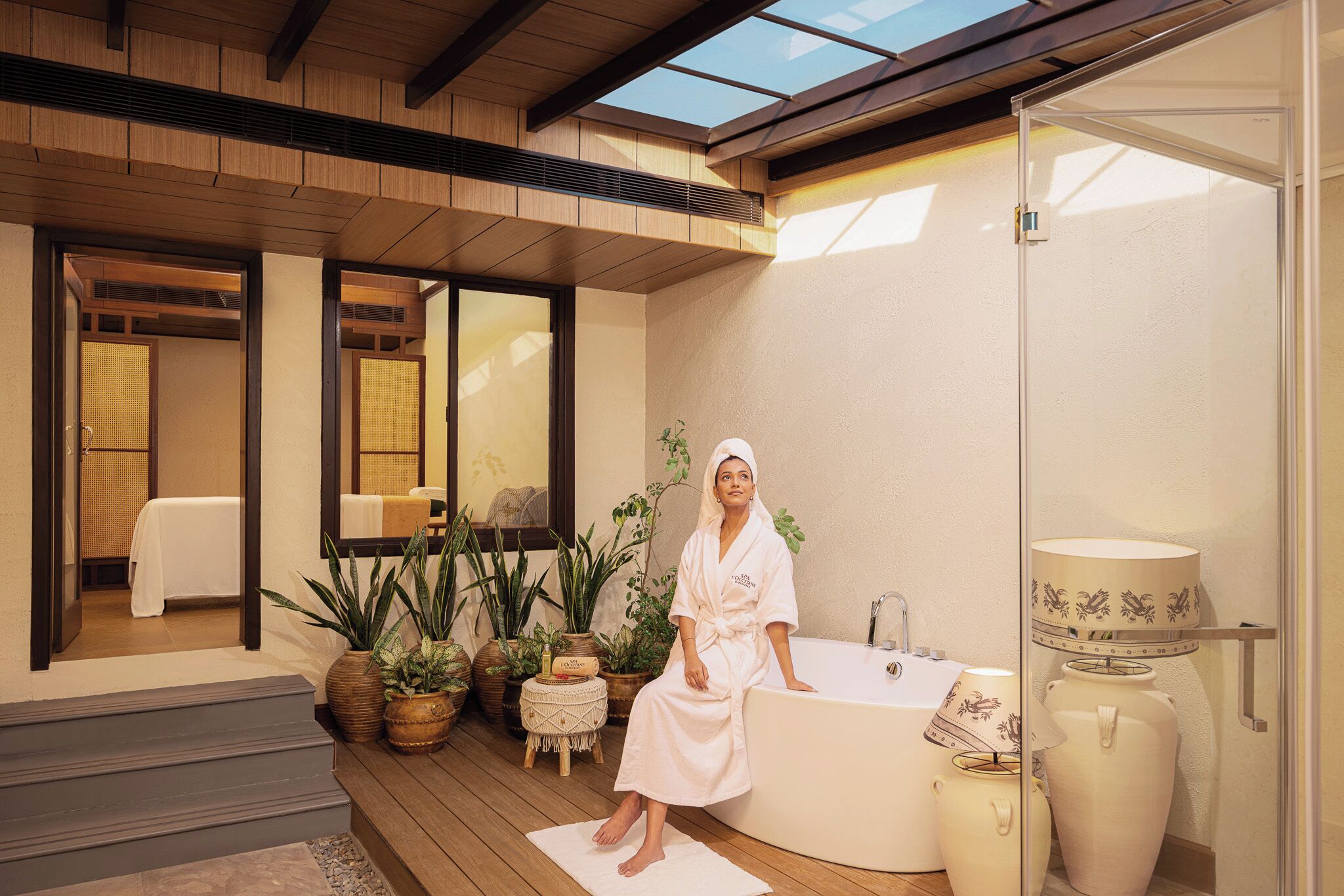 En person i badekåbe sidder i et badekar i et spa-lignende miljø med planter og et stort spejl.