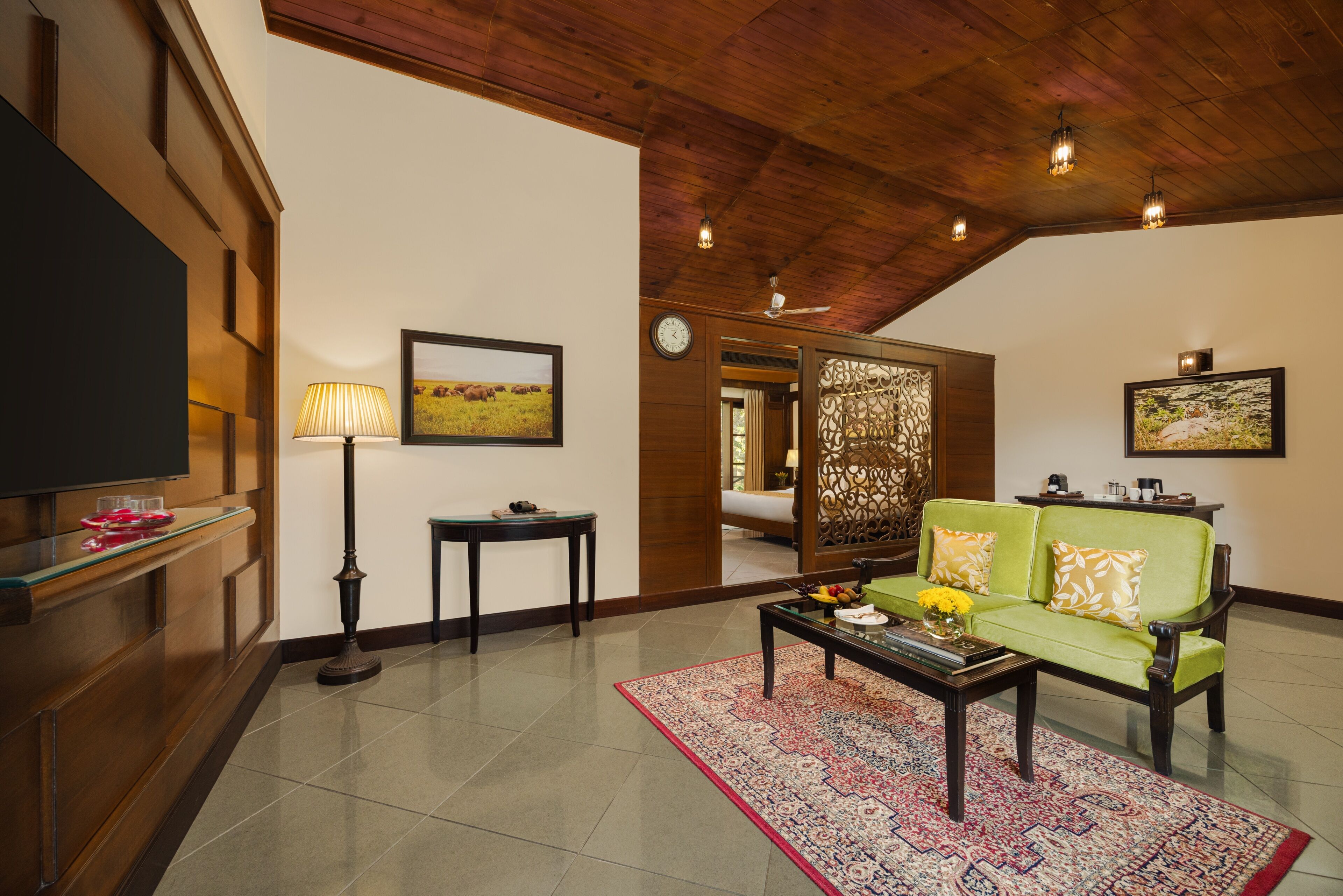 A spacious living room with a wooden ceiling, a green sofa, a coffee table with flowers, a TV, and a decorative screen.