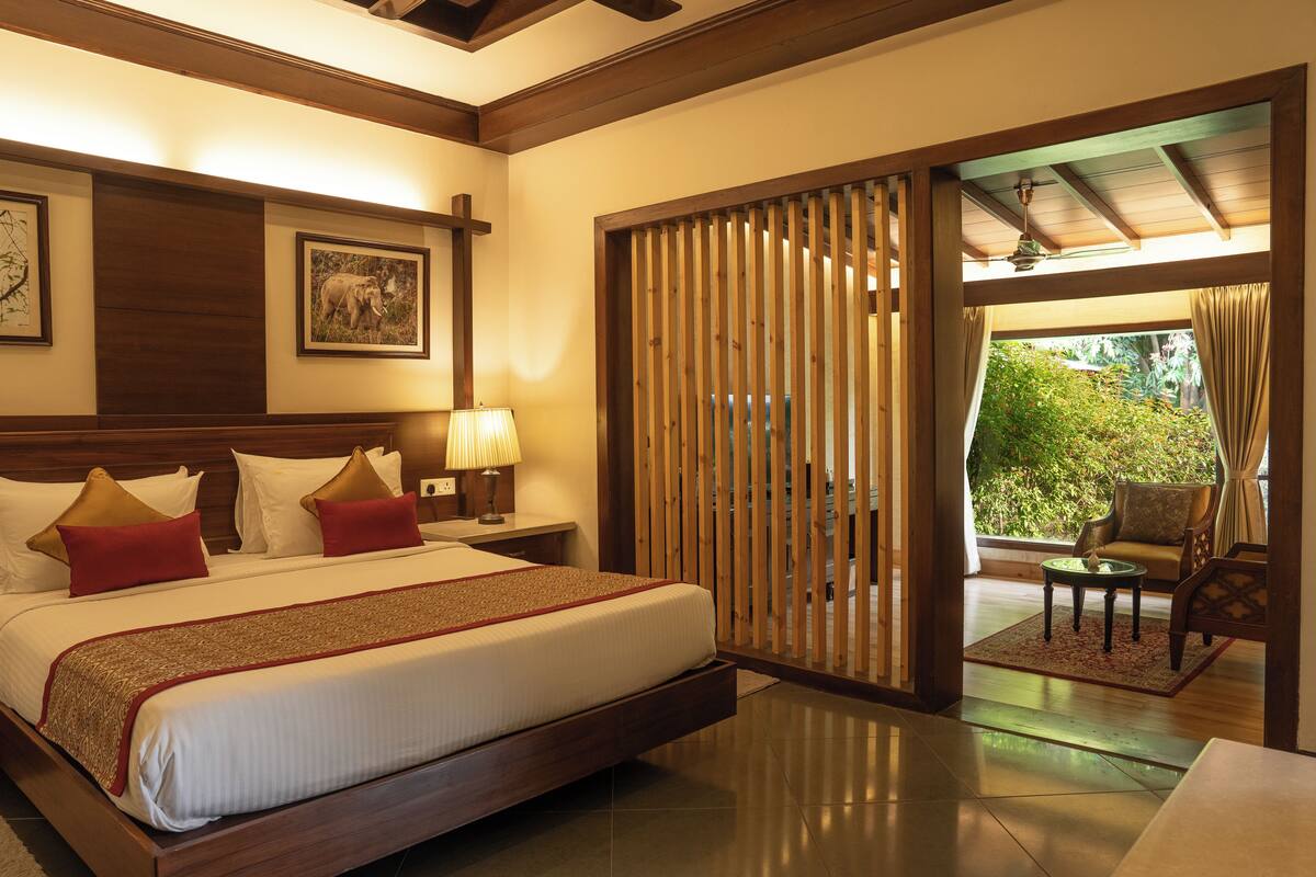 A hotel room with a large bed, wooden headboard, a nightstand, a lamp, and a view of the outdoors through a sliding door.