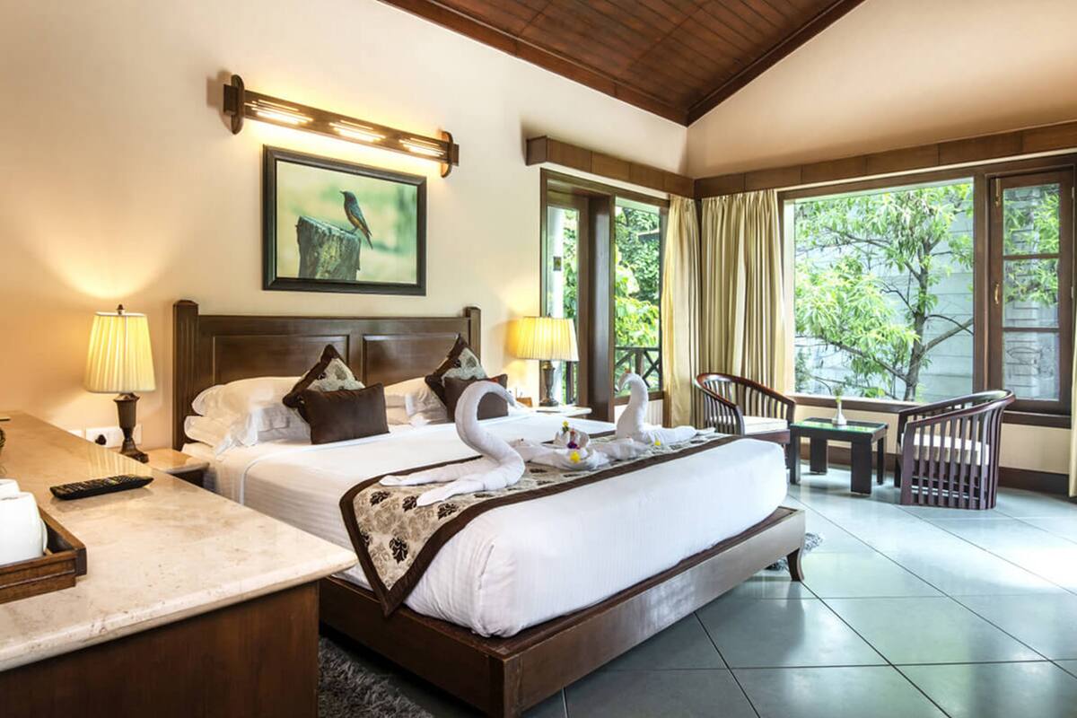 A hotel room with a large bed, two bedside tables, a desk, and a chair. There is a window with curtains and a view of greenery outside.