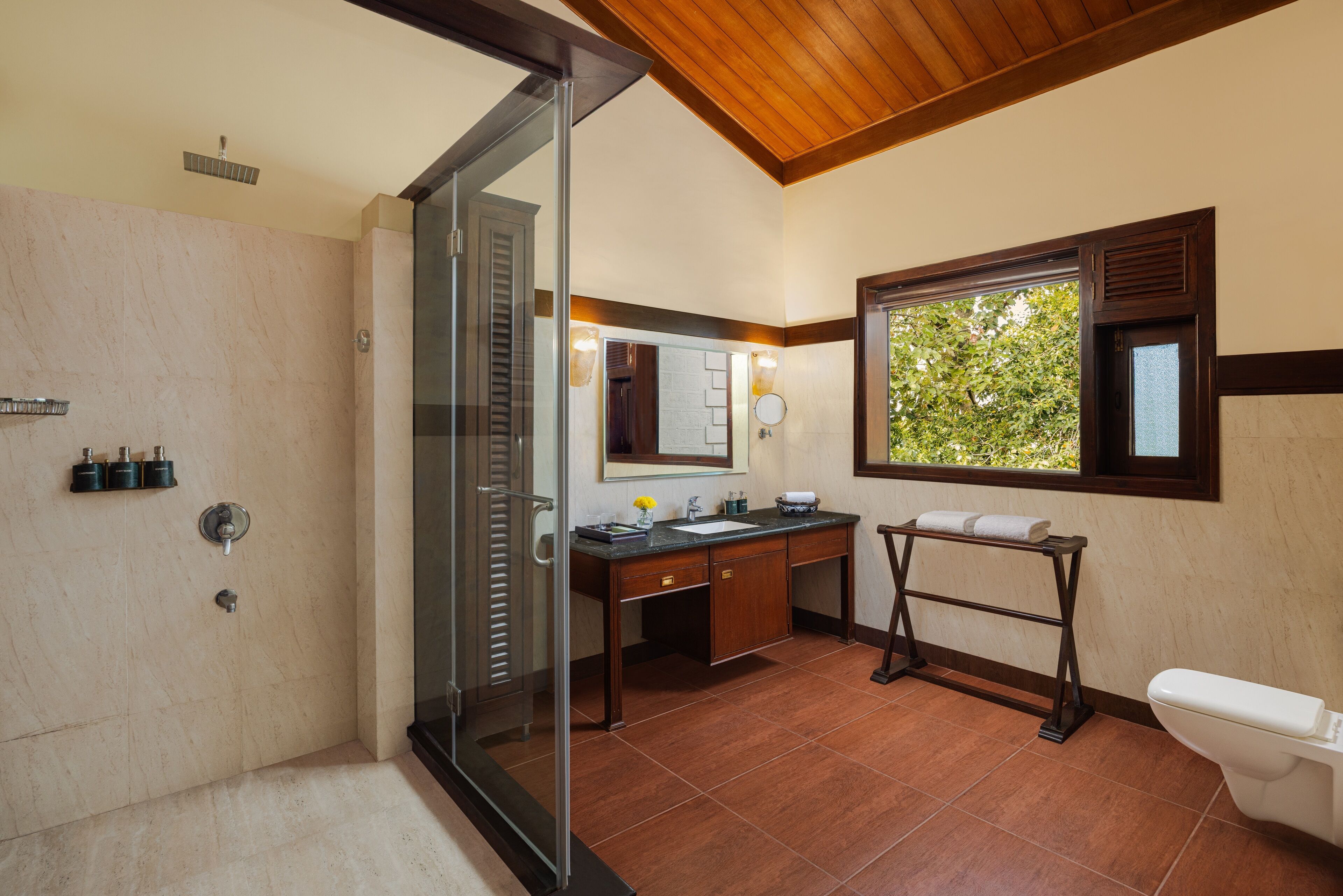 Un bagno con porta doccia in vetro, mobile lavabo in legno, lavandino, WC e finestra con vista sul verde.