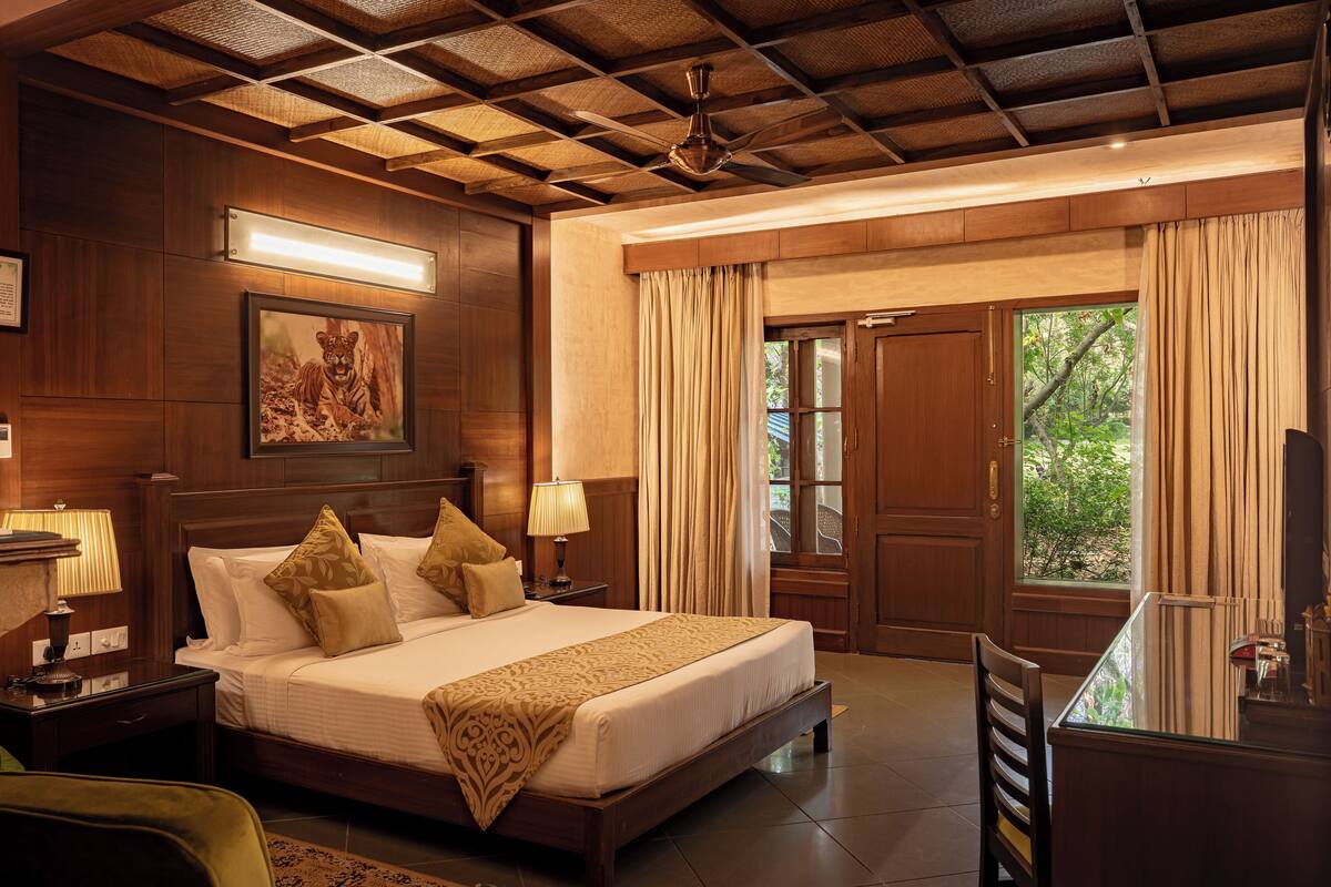 A hotel room with a large bed, wooden headboard, a ceiling fan, a nightstand with a lamp, a desk with a chair, and a window with curtains.