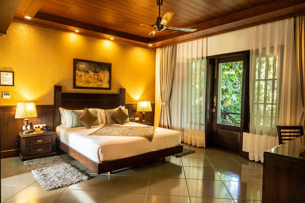 A well-lit bedroom with a large bed, wooden headboard, and a ceiling fan. There is a nightstand with a lamp and a framed picture on the wall.