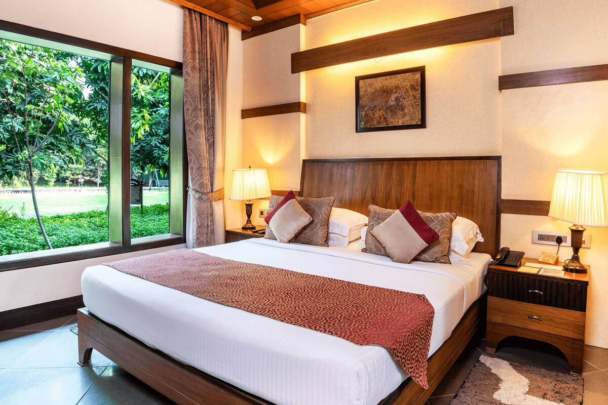 A hotel room with a large bed, two bedside tables with lamps, a window with curtains, a framed picture on the wall, and a view of greenery outside.