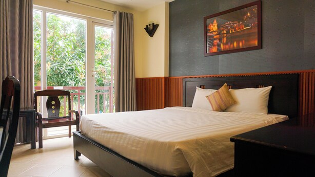 A hotel room with a large bed, a wooden headboard, a nightstand, a chair, and a view of greenery through the window. - Hoi An Paradise Hotel (Da Nang)