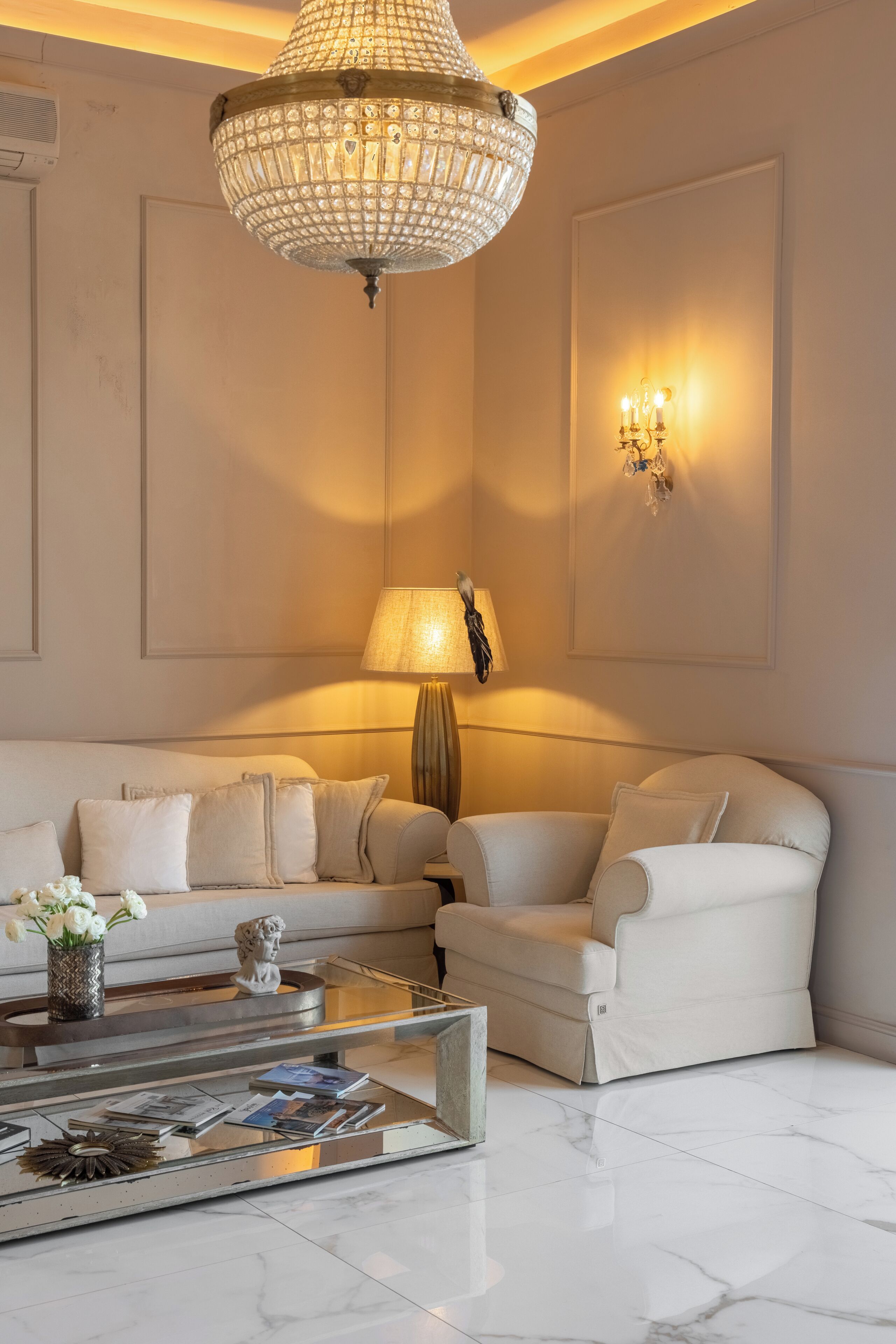 Un salon moderne avec un canapé blanc, une table basse en verre, un lustre en cristal et une lampe fixée au mur.
